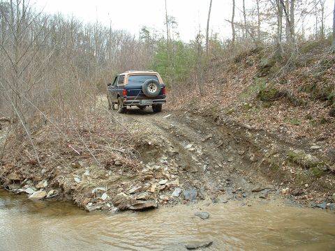 1987 Ford Bronco - A 1987 Ford Bronco navigates a challenging off-road path, with a muddy stream to its left.