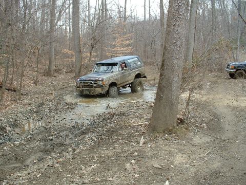1987 Ford Bronco - A 1987 Ford Bronco navigates a challenging muddy off-road course, with a second vehicle partially visible on the right.