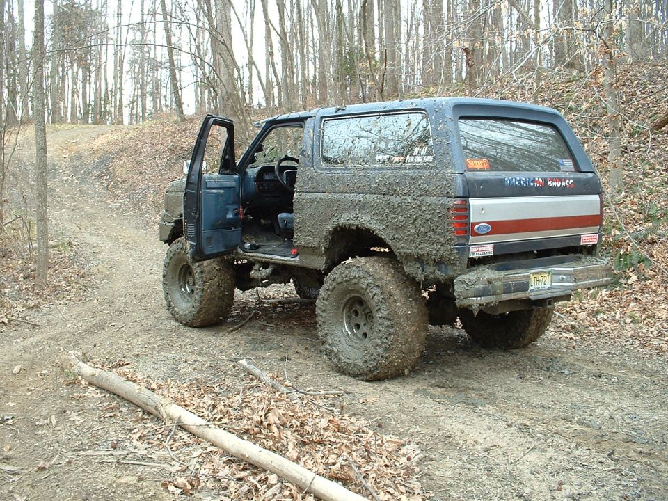 Muddy 1987 Ford Bronco with driver's door open, parked on a dirt trail in woods