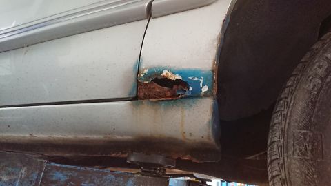1999 Ford Fairlane - Close-up of a corroded car panel, revealing extensive rust damage and a section of blue primer.