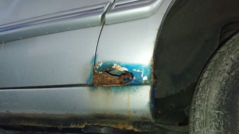 1999 Ford Fairlane - Significant rust damage is visible on the lower body panel of this vehicle, with blue undercoating showing through the corroded area.