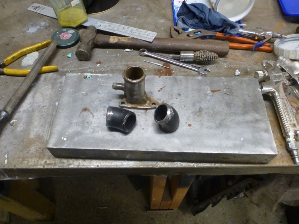 Metal pipe fittings and a thermostat housing sit on a flat metal plate. Tools like hammers, pliers, and a spray gun are on the workbench