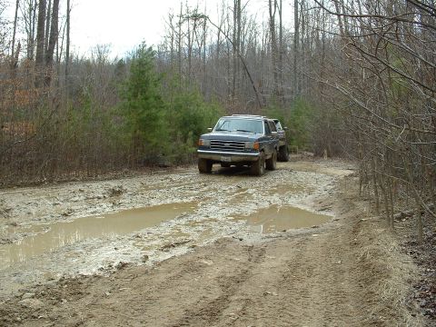 1987 Ford Bronco - A 1987 Ford Bronco navigates a challenging muddy path in a wooded area.