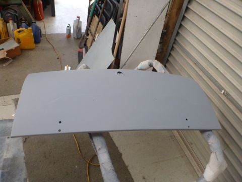 1962 MG B - A car trunk lid, prepped for paint, rests on supports in a workshop environment with garage.