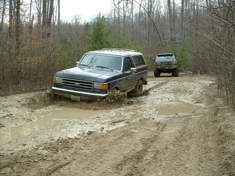 1987 Ford Bronco - Two SUVs navigate a muddy trail through a wooded area.