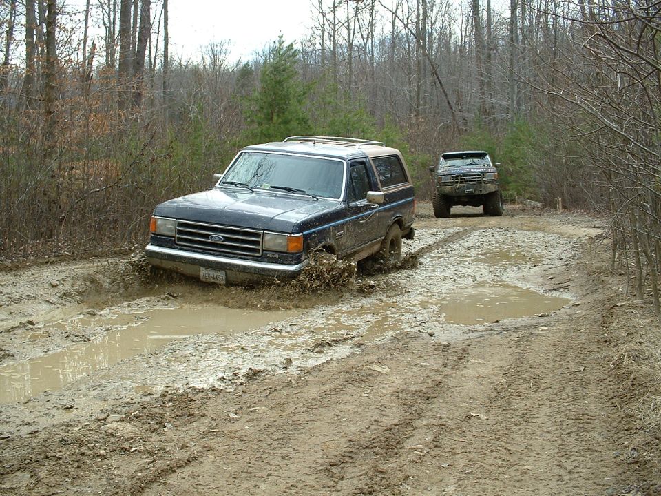 Dark blue Ford Bronco driving through mud, front quarter view. Another SUV follows behind