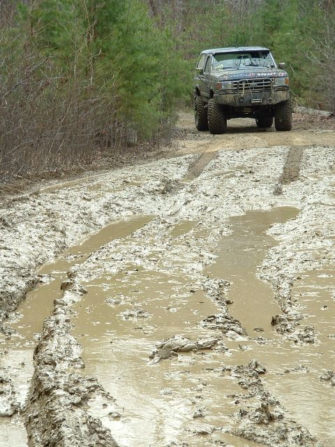 1987 Ford Bronco - A lifted Ford Bronco navigates a challenging muddy path in a wooded area.