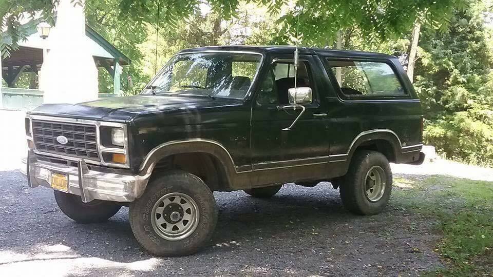 Black Ford Bronco parked on gravel with trees and a small building in the background