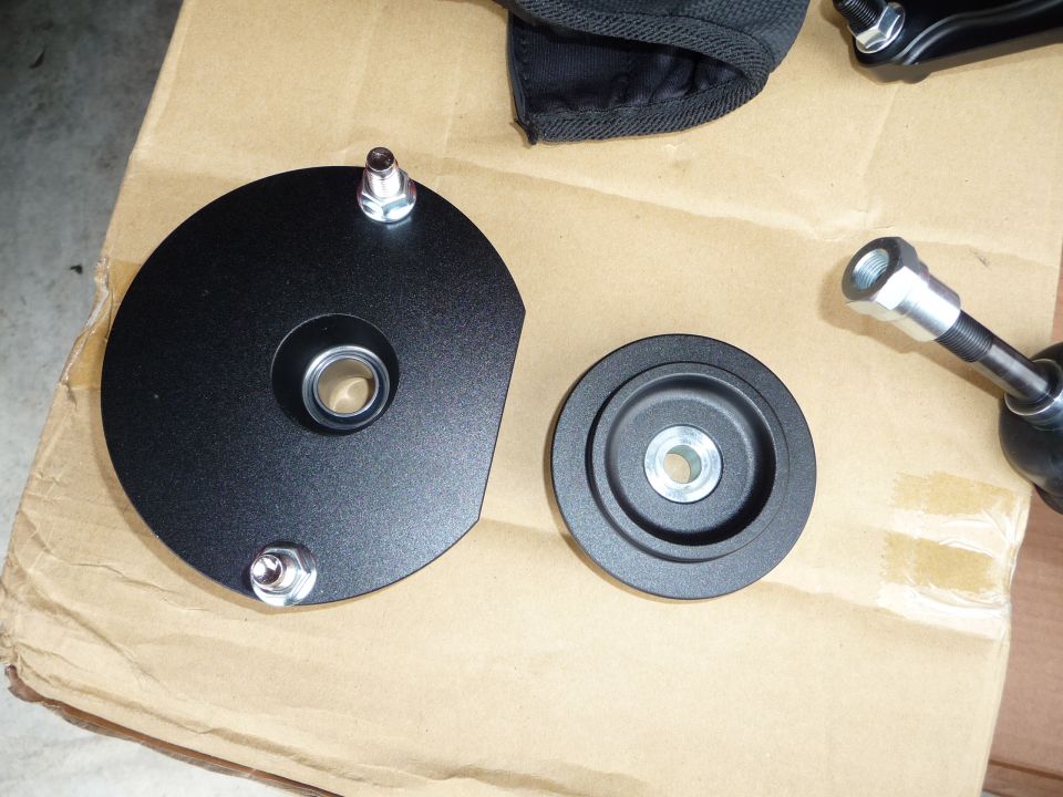 Two black metal suspension components with bolts and bearings on a cardboard surface