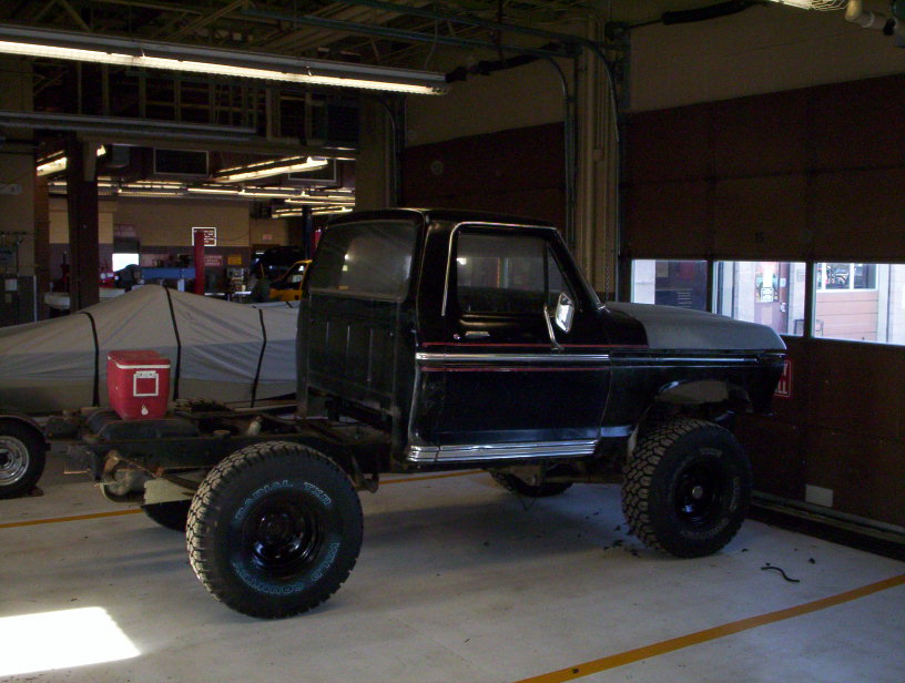 Black Ford F-Series truck chassis with a bare hood, viewed from the front-right in a garage