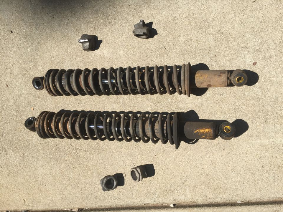 Two rusty shock absorbers with yellow bodies and coiled springs, along with four mounting hardware pieces, lie on a concrete surface