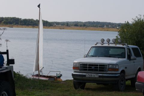 1993 Ford Bronco - A white Ford Bronco is parked on the grassy shore of a lake next to a small sailboat. The SUV has a light bar with four spotlights mounted…