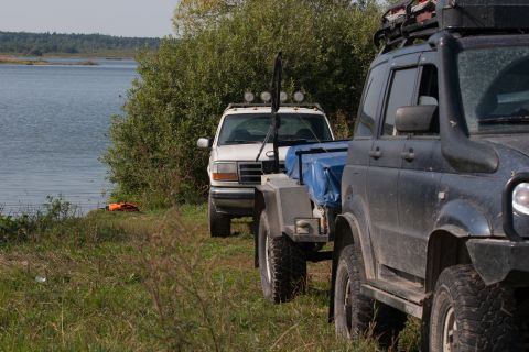 1993 Ford Bronco - A white Ford Bronco is parked by a lake, towing a trailer covered with a blue tarp. A dark SUV is parked in the foreground, obscuring part…
