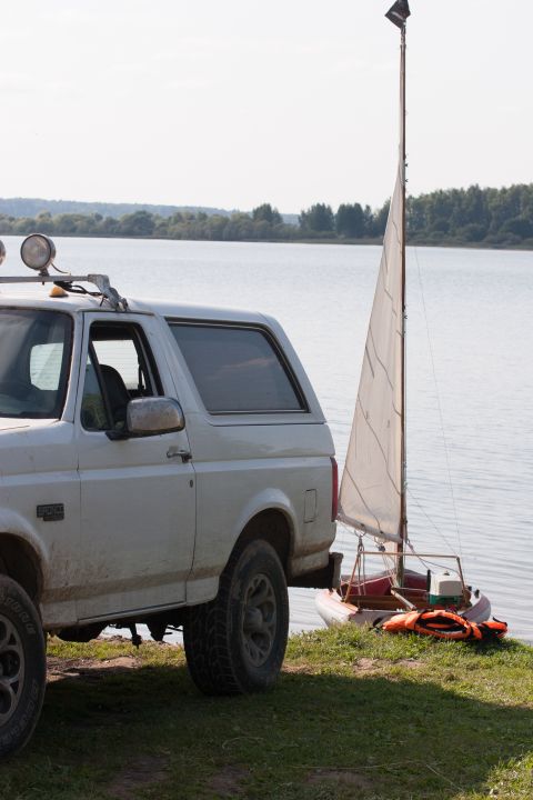1993 Ford Bronco - A white Ford Bronco is parked on the grassy shore of a lake next to a small sailboat with its sail up. The sailboat has an orange life vest…