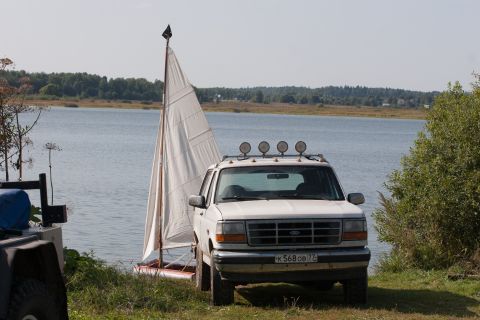 1993 Ford Bronco - A white Ford Bronco is parked next to a small sailboat on the grassy shore of a lake. The SUV has a roof rack with four lights and a licens…
