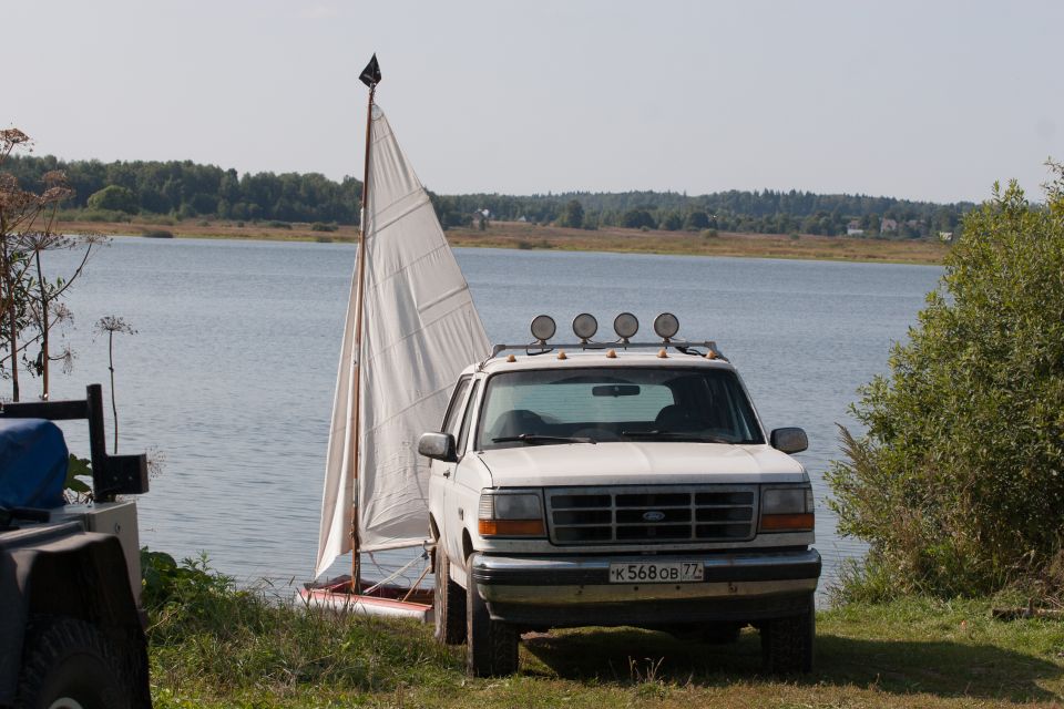 White Ford Bronco parked by a small sailboat near a lake.