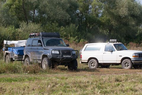 1993 Ford Bronco - A dark blue SUV with a roof rack and a trailer is parked next to a white Ford Bronco with auxiliary lights on the roof. Both vehicles are o…