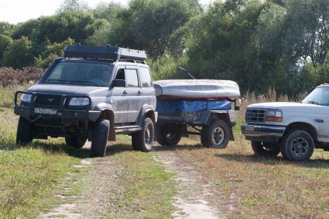 1993 Ford Bronco - A dark gray SUV tows a trailer with a boat on it, parked next to a white Ford Bronco on a dirt path with trees in the background.