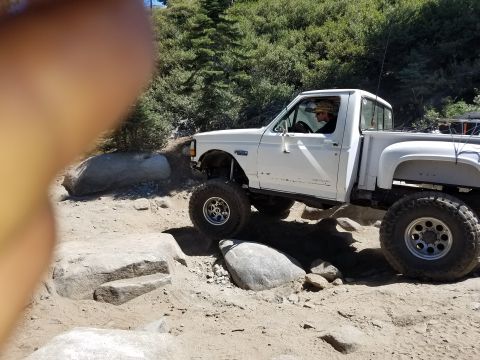 1968 Ford Bronco - A white, lifted pickup truck is navigating a rocky off-road trail. The driver is visible behind the wheel, wearing a hat.