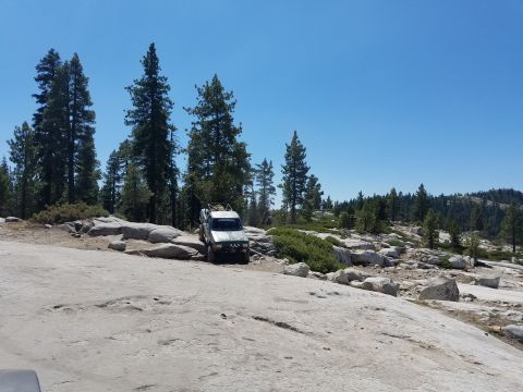 1968 Ford Bronco - A lifted, olive green SUV is parked on a rocky, mountainous terrain with scattered pine trees under a clear blue sky.