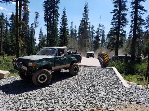 1968 Ford Bronco - A modified green off-road vehicle with large tires is parked on a gravel road leading to a bridge. Another white off-road vehicle is crossi…