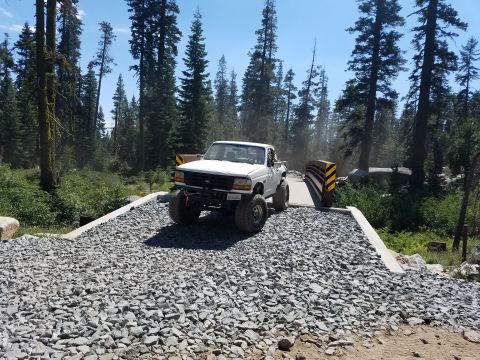 1968 Ford Bronco - A white lifted Ford pickup truck is driving across a gravel bridge in a forest setting. The truck has large off-road tires and a modified f…