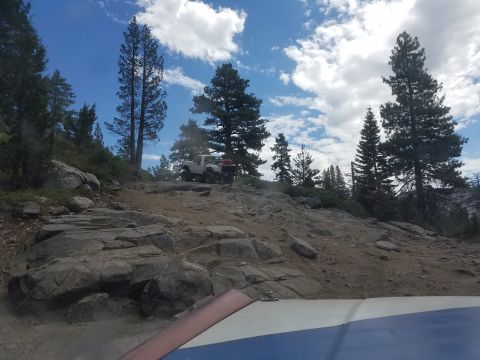 1968 Ford Bronco - A white pickup truck is parked on a rocky, uphill trail surrounded by pine trees under a partly cloudy blue sky. The front of another vehic…