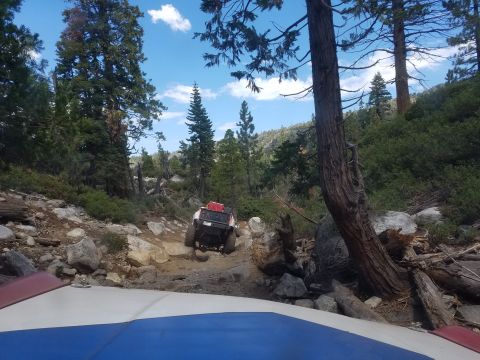 1968 Ford Bronco - A white off-road vehicle with red cargo boxes on top navigates a rocky, wooded trail. The view is from another vehicle, showing its hood in…