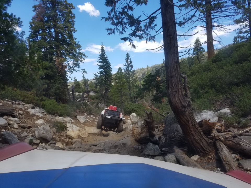 White off-road vehicle with red cargo on a rocky trail, seen from another vehicle's hood.