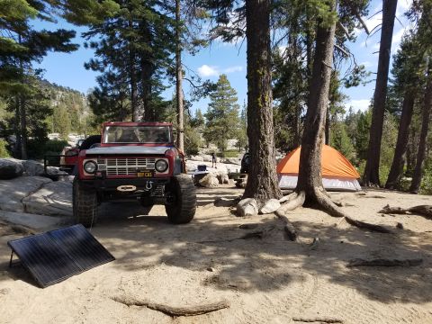 1968 Ford Bronco - A red 1968 Ford Bronco is parked on a dirt campsite next to an orange tent and a portable solar panel. The vehicle has large off-road tires…