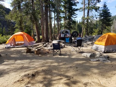 1968 Ford Bronco - A campsite is set up with two orange tents, camping chairs, and a red off-road vehicle parked amongst trees and rocks.