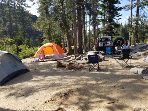 1968 Ford Bronco - A campsite scene features an orange and gray tent in the foreground, with a red off-road vehicle parked behind it. Several camping chairs a…
