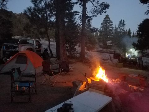 1968 Ford Bronco - A campsite scene unfolds at dusk, illuminated by a crackling campfire, with tents set up nearby.