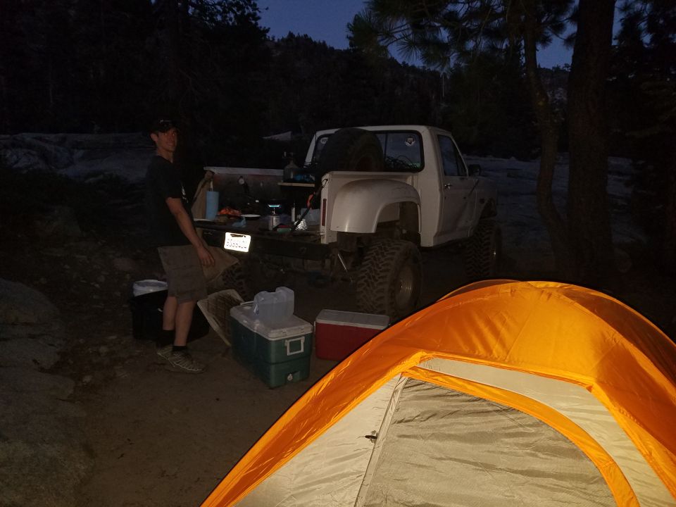 White pickup truck parked at a campsite with camping gear and a tent in the foreground. A person stands by the truck bed