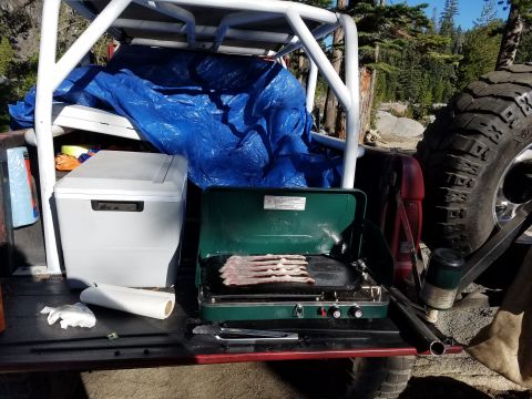 1968 Ford Bronco - Camping setup on a vehicle's tailgate with a cooler, stove cooking bacon, and a spare tire visible.
