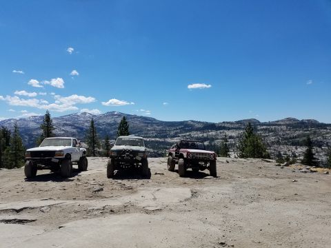 1968 Ford Bronco - Three modified off-road vehicles are parked on a rocky, elevated terrain with a mountainous, forested landscape in the background under a b…