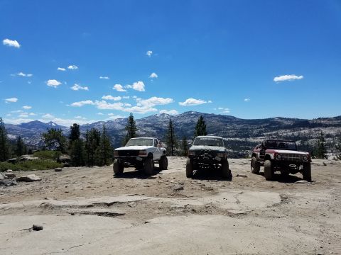 1968 Ford Bronco - Three lifted off-road vehicles are parked on a rocky, uneven terrain with a mountainous landscape and blue sky with scattered clouds in the…
