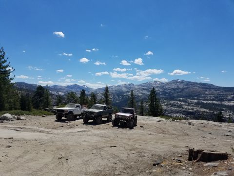 1968 Ford Bronco - Three off-road vehicles are parked on a rocky, dirt clearing with a mountainous, forested landscape in the background under a blue sky with…