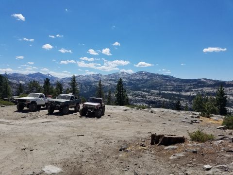 1968 Ford Bronco - Three modified off-road vehicles are parked on a rocky, dirt clearing with a vast mountain landscape and blue sky with scattered clouds in…