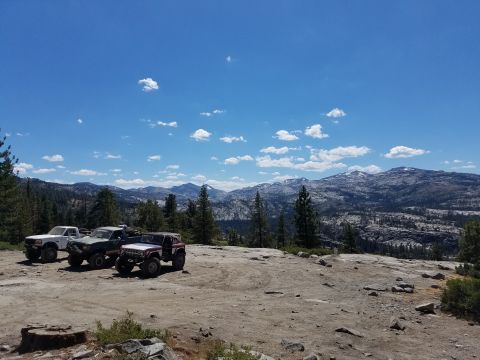 1968 Ford Bronco - Three off-road vehicles are parked on a rocky, dirt clearing with a mountainous, forested landscape and blue sky with scattered clouds in t…