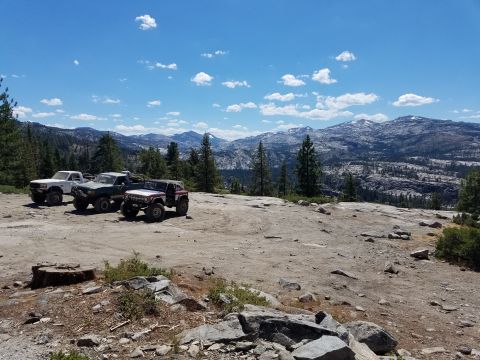 1968 Ford Bronco - Three off-road vehicles are parked on a rocky, elevated terrain with a mountainous landscape and blue sky with clouds in the background. Th…