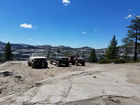 1968 Ford Bronco - Four off-road modified trucks are parked on a rocky, mountainous terrain under a clear blue sky with scattered clouds. The vehicles are pos…
