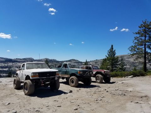 1968 Ford Bronco - Three modified off-road trucks are parked on a dirt road with a mountainous, tree-covered landscape in the background under a clear blue sk…