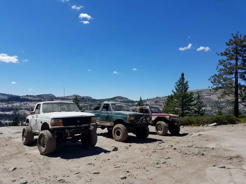 Three lifted off-road trucks parked on a dirt road with a scenic mountain backdrop.