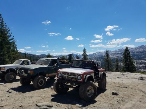 1968 Ford Bronco - Three off-road modified vehicles are parked on a dirt and rock surface with a mountainous, tree-filled landscape and a blue sky with scatte…