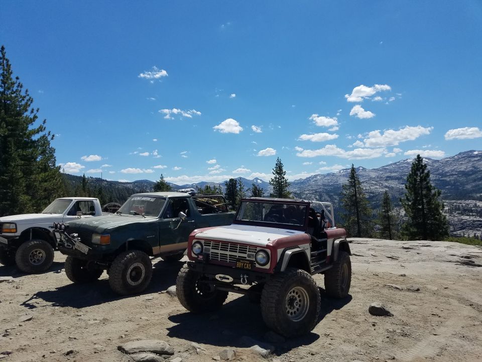 Three off-road vehicles parked on a rocky surface with mountains and trees in the background. The closest vehicle is a red and white Ford Bronco.