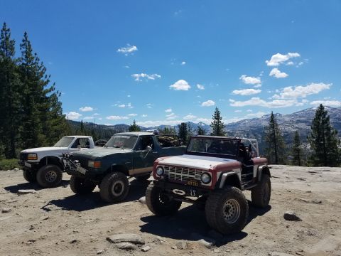 1968 Ford Bronco - Three modified off-road vehicles are parked on a rocky, dirt surface with a mountainous landscape and blue sky in the background. The vehic…