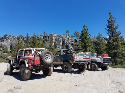 1968 Ford Bronco - Three modified off-road vehicles are parked on a dirt and rock surface with a rocky cliff and pine trees in the background under a clear bl…