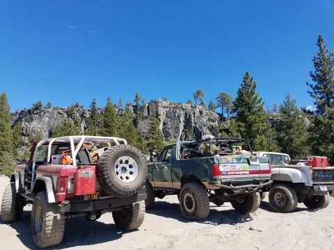1968 Ford Bronco - Three off-road modified trucks are parked on a dirt road with a rocky cliff face and pine trees in the background. The truck in the foregro…