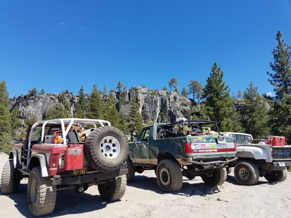Three off-road trucks parked with a rocky cliff and trees in the background. Red and white Ford Bronco in foreground.
